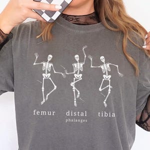 May include: Gray t-shirt featuring a white graphic of three dancing skeletons. The shirt has the words "femur, distal, tibia, phalanges" printed below the skeletons. The shirt has short sleeves and a crew neck.