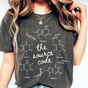 May include: Dark gray t-shirt with the text "the source code" and scientific diagrams. The shirt features the words Adenine, Uracil, Cytosine, Thymine, and Guanine. The shirt is a casual, unisex style.