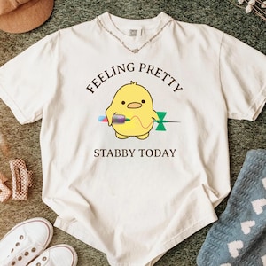 May include: Cream-colored t-shirt with the text "Feeling Pretty, Stabby Today" and a cartoon yellow chick holding a syringe. The shirt is surrounded by a brown hat, white sneakers, a silver necklace, and a blue patterned blanket.