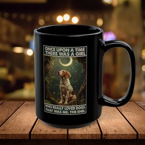 May include: Black ceramic mug with a dog illustration under a crescent moon and trees. The mug has the text "ONCE UPON A TIME THERE WAS A GIRL WHO REALLY LOVED DOGS THAT WAS ME. THE END." printed on it. The mug is on a wooden surface.