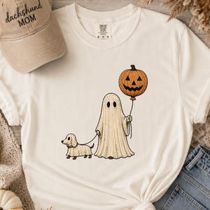 May include: A cream-colored t-shirt featuring a graphic of a ghost holding a pumpkin balloon and walking a dachshund dog in a ghost costume. Beside the shirt is a tan baseball cap with the embroidered text dachshund MOM on the front.
