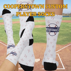 以下が含まれることがあります： 黒いかかととつま先が付いた白い野球用ソックス。野球のデザインと「COOPERSTOWN CUSTOM PLAYER SOCKS」の文字が特徴です。ソックスには野球のグラフィックと「COOPERSTOWN NAME ## 2026」の文字があります。