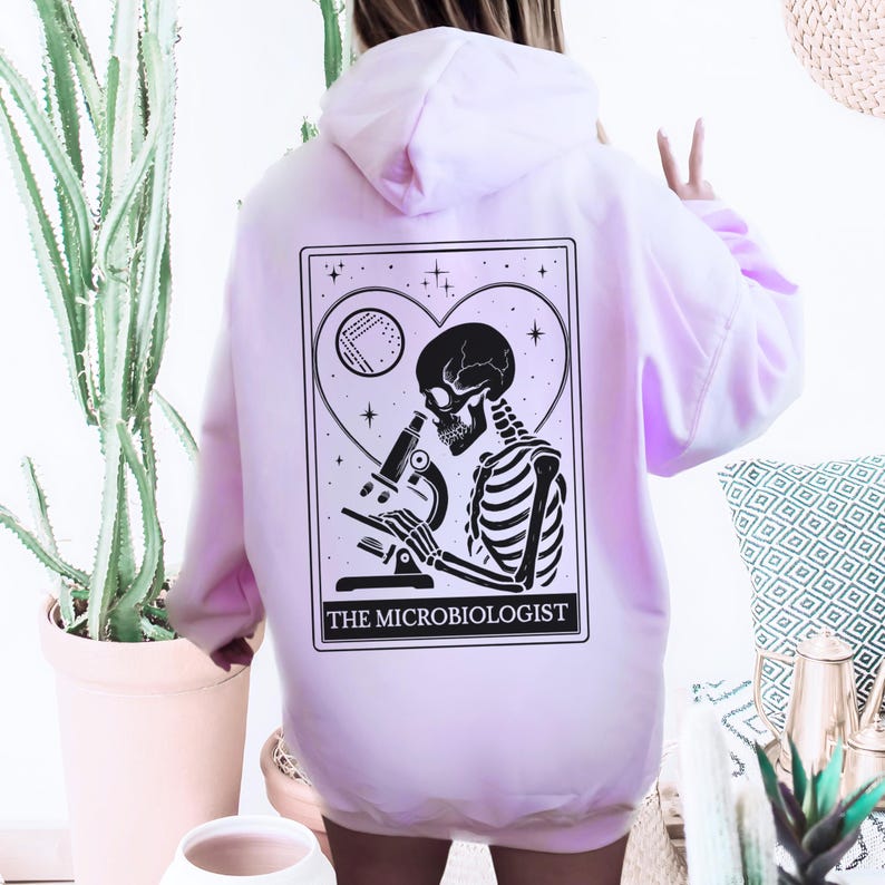May include: Light pink hoodie with a black and white graphic of a skeleton using a microscope, inside a heart-shaped frame. The text "THE MICROBIOLOGIST" is below the graphic. The hoodie has a hood and long sleeves.