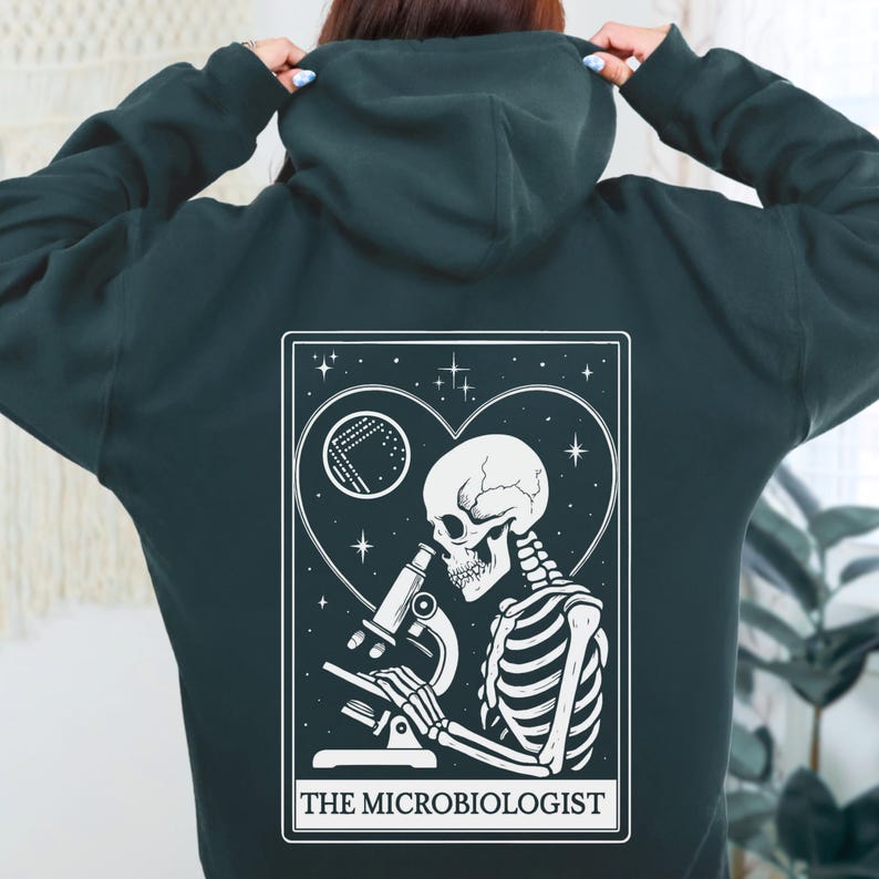 May include: Dark teal hoodie with a white graphic on the back. The graphic features a skeleton looking through a microscope, inside a heart-shaped frame with stars and a moon. Text at the bottom reads "THE MICROBIOLOGIST".