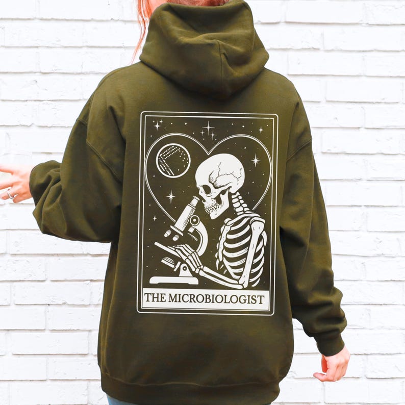 May include: Olive green hoodie with a white graphic of a skeleton using a microscope, set within a heart-shaped frame. The text "THE MICROBIOLOGIST" is printed below the graphic. The hoodie has a hood and long sleeves.