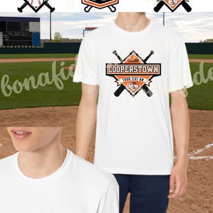 Puede incluir: Camiseta blanca con un diseño de béisbol, con el texto "COOPERSTOWN" y "2026". El diseño incluye bates de béisbol, una pelota de béisbol y una forma de escudo. La camiseta la lleva una persona.