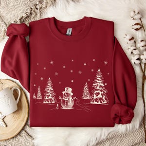 May include: A dark red crewneck sweatshirt featuring a white graphic print of a central snowman wearing a top hat and scarf, surrounded by several pine trees and falling snowflakes. The garment has long sleeves and a ribbed collar and cuffs.