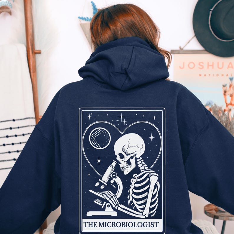 May include: Navy blue hoodie featuring a white graphic of a skeleton using a microscope, set within a heart and star design. The text "THE MICROBIOLOGIST" is at the bottom. The hoodie has a drawstring hood and a kangaroo pocket.
