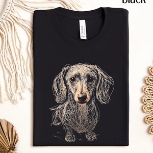 May include: Black t-shirt featuring a sketched dachshund dog design. The dog is depicted in a detailed, artistic style with a light brown and white color palette. The word "black" is printed at the top right corner.