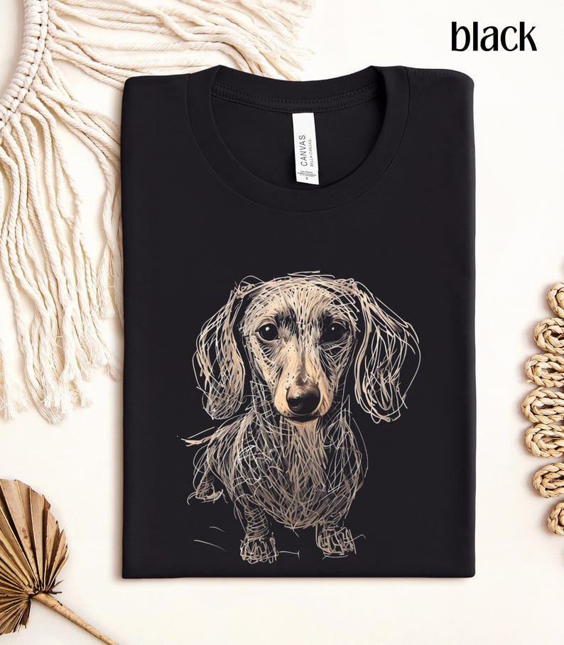 May include: Black t-shirt featuring a sketched dachshund dog design. The dog is depicted in a detailed, artistic style with a light brown and white color palette. The word "black" is printed at the top right corner.