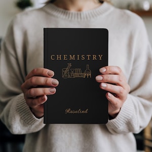 May include: A black notebook featuring the word "CHEMISTRY" in gold and gold illustrations of laboratory glassware. The name "Rosalind" is printed in gold script below the illustrations.