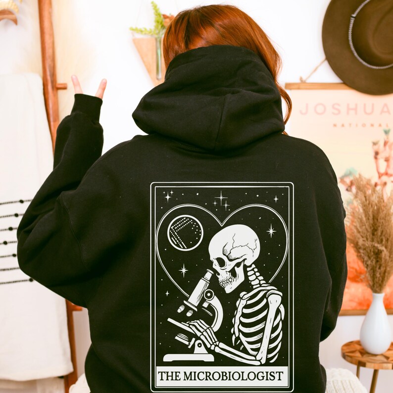 May include: Black hoodie featuring a white graphic of a skeleton using a microscope, set within a heart-shaped frame with stars and a petri dish. The text "THE MICROBIOLOGIST" is at the bottom of the design. The hoodie has a hood and long sleeves.