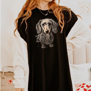 May include: Black t-shirt featuring a detailed sketch of a dachshund dog. The word "black" is printed in the upper right corner. The dog illustration is in a line art style, with fine details. The shirt is a casual style.