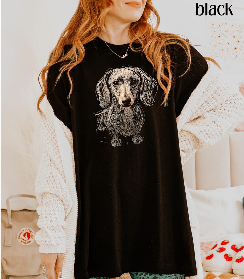 May include: Black t-shirt featuring a detailed sketch of a dachshund dog. The word "black" is printed in the upper right corner. The dog illustration is in a line art style, with fine details. The shirt is a casual style.