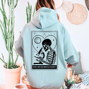 May include: A light blue hoodie featuring a black and white graphic of a skeleton using a microscope, set within a heart-shaped design. The text "THE MICROBIOLOGIST" is printed below the graphic. The hoodie is made of a soft material.