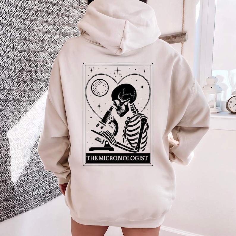 May include: Cream-colored hoodie featuring a black and white graphic of a skeleton using a microscope, set within a heart-shaped frame. The text "THE MICROBIOLOGIST" is printed below the graphic. The hoodie has a hood and a kangaroo pocket.