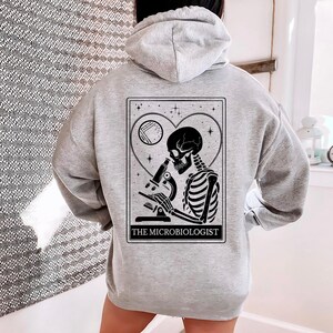 May include: A light gray hoodie featuring a black and white graphic of a skeleton using a microscope, set within a heart-shaped frame. The text "THE MICROBIOLOGIST" is printed below the image. The hoodie has a drawstring hood.