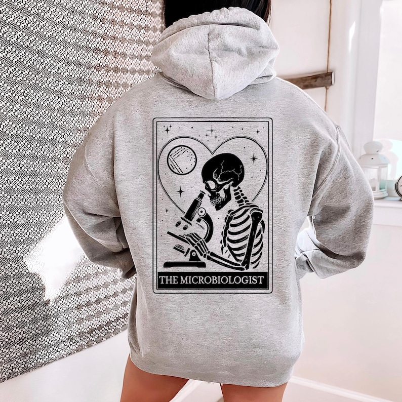 May include: A light gray hoodie featuring a black and white graphic of a skeleton using a microscope, set within a heart-shaped frame. The text "THE MICROBIOLOGIST" is printed below the image. The hoodie has a drawstring hood.