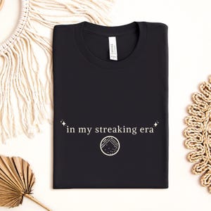 May include: Black t-shirt with the text "in my streaking era" in white, along with a circular graphic. The shirt is folded neatly. The shirt is made of a soft material.