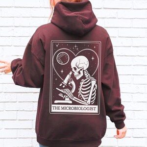 May include: Burgundy hoodie with a white graphic on the back. The graphic features a skeleton looking through a microscope, inside a heart-shaped frame, with the text "THE MICROBIOLOGIST". The hoodie has a hood and long sleeves.