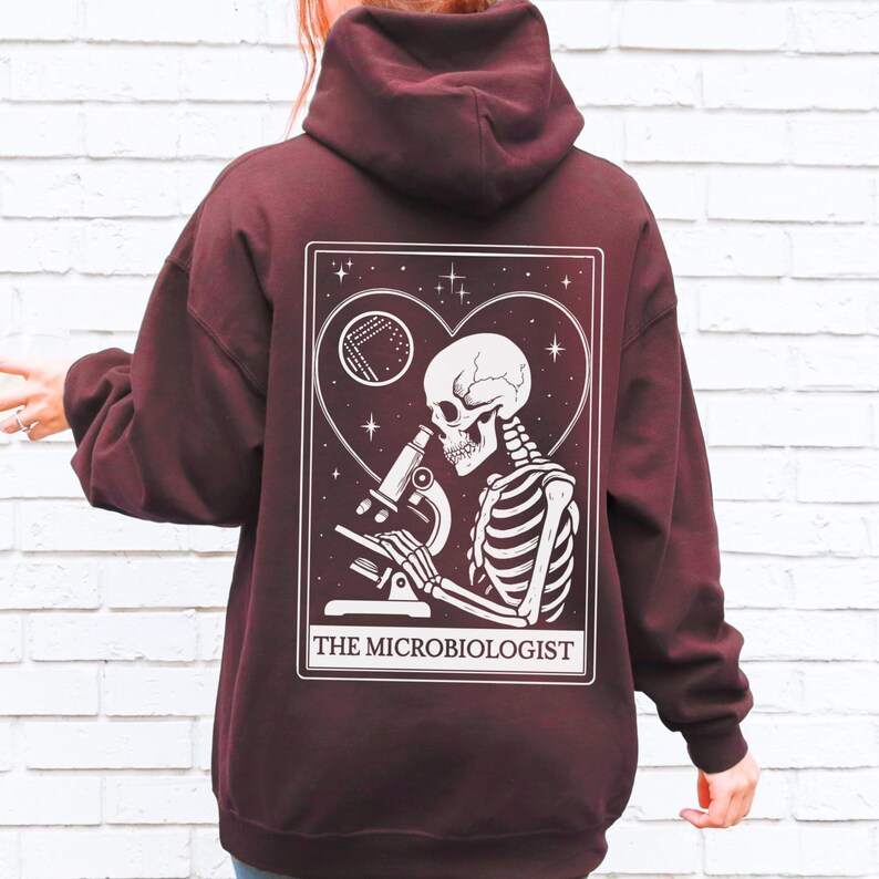 May include: Burgundy hoodie with a white graphic on the back. The graphic features a skeleton looking through a microscope, inside a heart-shaped frame, with the text "THE MICROBIOLOGIST". The hoodie has a hood and long sleeves.
