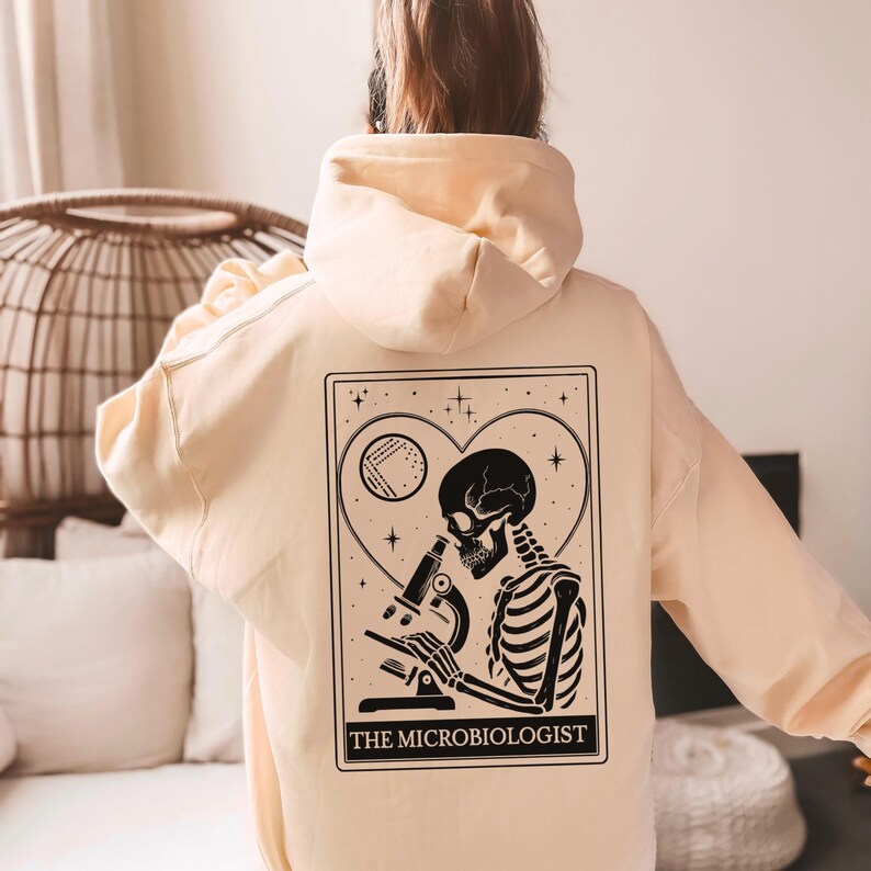 May include: A light peach-colored hoodie with a black graphic on the back. The graphic features a skeleton using a microscope, inside a heart shape, with the text "THE MICROBIOLOGIST". The hoodie has a hood and long sleeves.