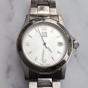 Puede incluir: Un reloj ESQ Swiss plateado con esfera blanca. El reloj tiene una pantalla de fecha a las 3 en punto y una correa plateada. La esfera del reloj tiene números negros en las posiciones de las 12, 6 y 9.