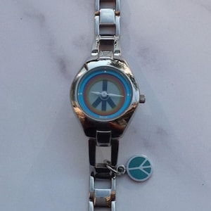 May include: Silver-toned wristwatch with a peace sign on the face. The watch has a rainbow-colored inner dial and a small charm with a peace symbol. The band is made of linked metal segments.