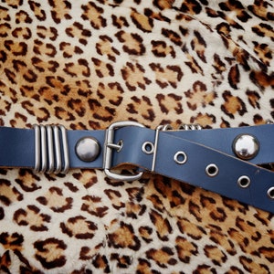 May include: A dark blue leather belt with a silver buckle and multiple silver rings. The belt is lying on a leopard print fabric.