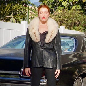 Könnte beinhalten: Schwarze Lederjacke mit cremefarbenem Pelzkragen. Die Jacke ist geknöpft und wird über einem schwarzen Oberteil getragen. Die Person steht vor einem schwarzen Auto.