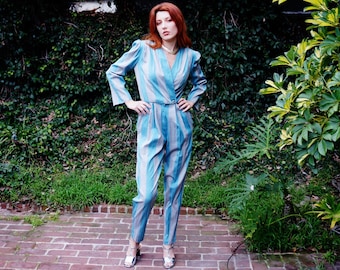May include: A blue and gray striped jumpsuit with a wrap-style top and a matching belt. The jumpsuit has long sleeves and is paired with silver heels. The model is standing on a brick patio with greenery in the background.