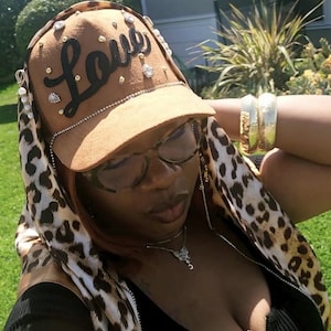 May include: A tan baseball cap featuring the word Love in black script lettering, embellished with scattered clear rhinestones and small gold studs. The cap includes a leopard print fabric attachment that drapes down the sides.