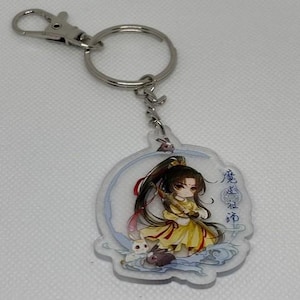 May include: A clear acrylic keychain featuring a cartoon character in a yellow dress with long black hair. The keychain has a silver clasp and a ring. The character is surrounded by rabbits and the text "魔道祖师".