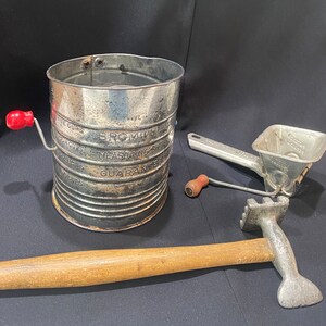 May include: Vintage silver-tone kitchen tools, including a flour sifter with a red handle and a hand-crank potato ricer. The sifter has the words "Bromwell Measuring Guarantee" printed on the side.