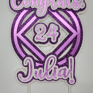May include: A purple glitter cake topper with the words "Congrats Julia!" and the number "24". The design features a layered effect with white and purple accents, mounted on white sticks.