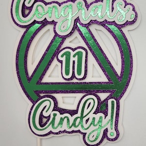 May include: A cake topper with the words "Congrats Cindy!" in green glitter script. The number "11" is in the center of a green and purple glitter triangle. The topper is on a white background.