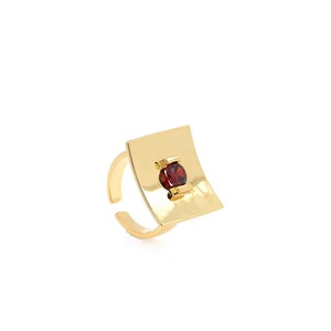 May include: A gold-coloured adjustable ring with a square face and a hammered texture. A small, round, red gemstone is set in the centre of the square. The ring has a simple, elegant design.