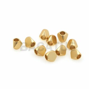 Puede incluir: Una colección de cuentas geométricas doradas. Cada cuenta tiene una forma facetada única con un agujero en el centro. Las cuentas están dispersas sobre un fondo blanco, mostrando su brillo metálico. Ideal para la fabricación de joyas.