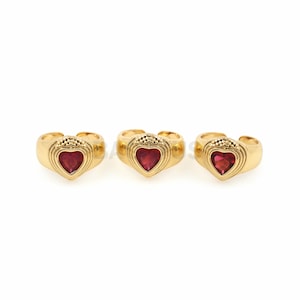 May include: Three gold rings with red heart-shaped gemstones. The rings are designed with a textured band and a heart-shaped setting.