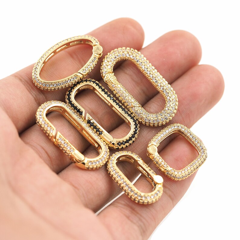 May include: Five gold-toned oval-shaped clasps with clear and black cubic zirconia stones. The clasps are on a hand.
