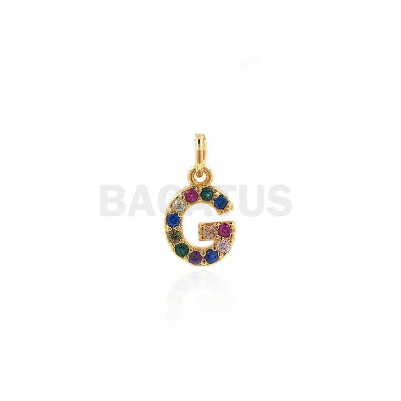 Puede incluir: Encanto de tono dorado con la letra G, adornado con piedras preciosas de colores.