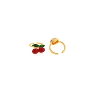 Pu&ograve; includere: Due anelli regolabili color oro con disegni di ciliegie. Un anello presenta un paio di ciliegie rosse con foglie verdi. L'altro anello ha una singola ciliegia rossa. Gli anelli sono su sfondo bianco.