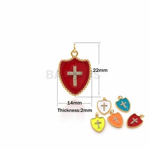 Pode incluir: Charme de pingente em forma de escudo em tom dourado com acabamento em esmalte vermelho e uma cruz de cristal transparente. O charme mede 14 mm de largura e 22 mm de altura. O pingente tem 2 mm de espessura. A imagem também mostra o charme em outras cores: branco, turquesa, amarelo, laranja e vermelho.