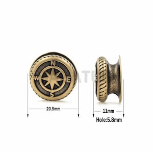 Puede incluir: Un amuleto de brújula de metal dorado con un diseño de cuerda alrededor del borde. La brújula presenta una estrella con las letras N, S, E y W alrededor. El amuleto mide 20,5 mm de diámetro y tiene un agujero de 5,8 mm.