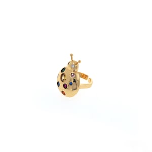 Peut inclure: Une bague dorée en forme de coccinelle avec des accents de pierres précieuses colorées. La bague présente un corps rond avec de multiples gemmes colorées et un petit diamant. La bague a une finition lisse et polie.