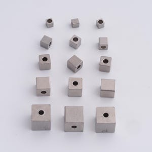 Pode incluir: Uma variedade de pequenas contas metálicas quadradas com um acabamento fosco. Cada conta tem um furo perfurado no centro e variam ligeiramente de tamanho. As contas estão dispostas sobre uma superfície branca.