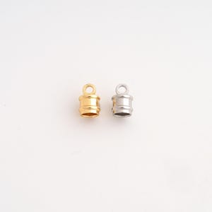 May include: Two cylindrical cord end caps for jewelry making, one finished in a polished gold tone and the other in a polished silver tone. Each cap features a smooth, rounded body with a small circular loop at the top for attachment purposes.