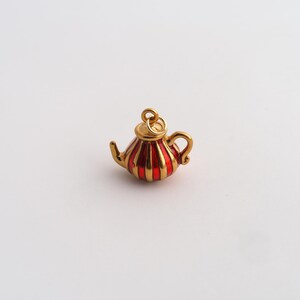 May include: A small gold-toned metal charm shaped like a teapot, featuring vertical red enamel stripes along the body, a curved handle, a spout, and a circular jump ring attached to the top for hanging as a piece of jewelry or accessory.