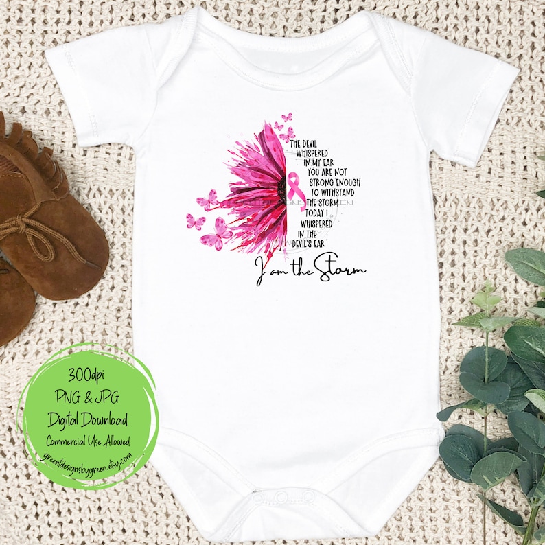 May include: A white baby onesie with a pink watercolor sunflower and a pink ribbon. The text reads "The devil whispered in my ear you are not strong enough to withstand the storm. Today I whispered in the devil's ear I am the storm."