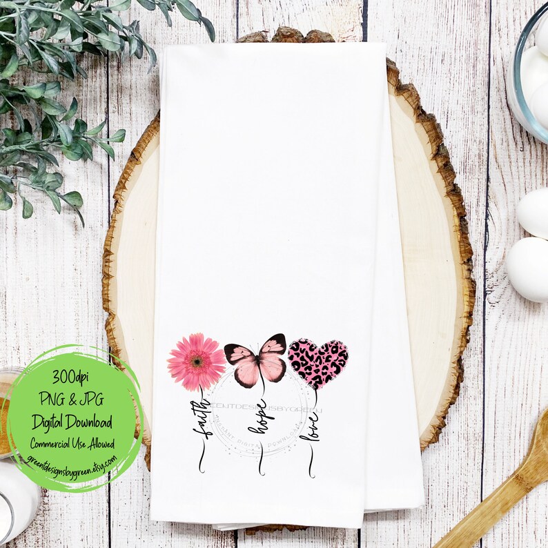 May include: White kitchen towel with a pink flower, butterfly, and heart with a leopard print design. The words "faith", "hope", and "love" are written around the design.